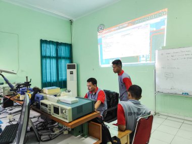 Makassar, 5 Nisan 2024 - Sınıfta bilgisayar ve laboratuvar ekipmanlarıyla elektronik öğrenen öğrenciler, arka planda projektör ekranı ve beyaz tahta.