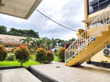 Sarı duvarları ve beyaz merdivenleri olan bir okul ya da ofis manzarası. Arka planda yemyeşil ağaçlar, kırmızımsı yapraklarla süslenmiş süs bitkileri ve paslı çatısı olan bir başka uzun bina var..