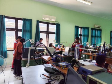 Makassar, 5 Nisan 2024 - Bir elektronik laboratuvarında çalışan öğrenciler, yeşil duvarları ve mavi perdeleri olan bir sınıfta çeşitli aletler ve makineler kullanıyorlar..