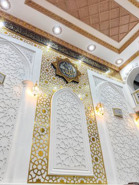 Wajo, 10 Nisan 2024 - Caminin karmaşık duvarları, altın İslami geometrik desenlerle çevrili ve sıcak ortam ışıklarıyla aydınlatılmış Arap kaligrafisinde Allah sözcüğünü içerir..