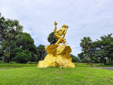 Bir kaya kaidesi üzerinde dans eden bir çiftin yan açılı görüntüsü. Arka planda palmiye ağaçları ve bulutlu gökyüzü olan yeşil bir kavşakta geçiyor..