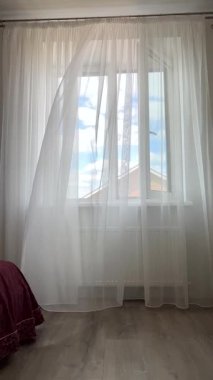 Açık bir yaz gününde rüzgâr tülleri açık pencerede uçuşur. Gökyüzü evin penceresinden görülebilir..