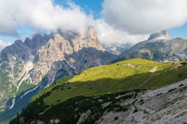 Güneşli yaz gününde Dolomitlerdeki dağ zirvelerinin önü yeşil çayır, arkaplanda bulutlar ve mavi gökyüzü tepede..