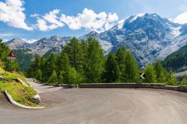 Sharp hairpin turn leads up the grassy mountain in the stunning sunlit Dolomites. Picturesque view of the snow capped mountain range above the winding asphalt road high in the Italian mountains.