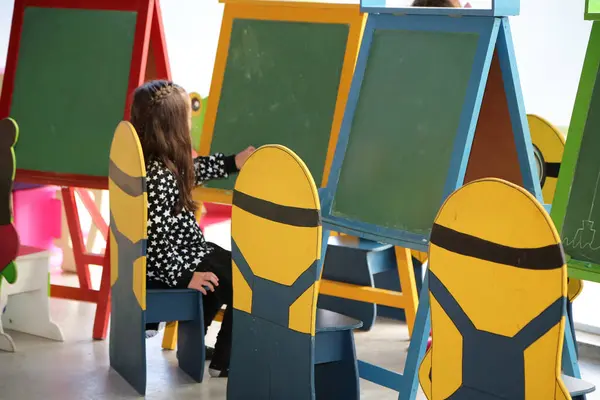 stock image Little girl drawing with chalks on green blackboard in a kindergarten. Child in initial level school. Little girl sitting on wooden benches shaped like minions.