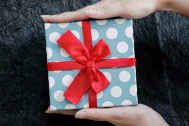 Hediye kutusunu tutan kadının elleri. Parlak kırmızı düğümlü fiyonk ve noktalı ambalaj kağıdı ve kurdeleyle süslenmiş hediye kutusu. Soyut arkaplan. Hediye, kutlama, yıldönümü konsepti. 