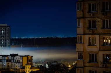 Nighttime cityscape with illuminated buildings in the foreground. A tall apartment building with glowing windows, a misty fog partially obscuring the view, creating a dramatic contrast. Kyiv, Ukraine clipart