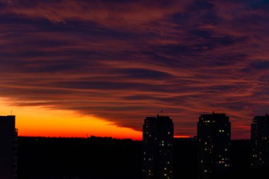 İki uzun binanın silueti dramatik turuncu-kırmızı günbatımı gökyüzüne karşı bulutların çizgileriyle. Görüntünün alt kısmı karanlık, günbatımının yansımaları bir su kütlesi üzerinde görünür..