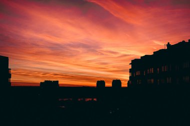 Yerleşim yerlerinin silueti dramatik turuncu-kırmızı günbatımı gökyüzüne karşı bulutların çizgileriyle. Görüntünün alt kısmı karanlık, günbatımının yansımaları bir su kütlesi üzerinde görünür..