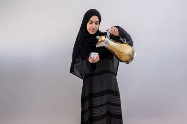 Sıcaklık ve gelenek saçan Arap bir kadın, ramazan ve bayram boyunca gösterilen konukseverliğin ve sevincin sembolü olan el üstünde tutulan Arap kahve fincanını, dingin, beyaz bir arka plana nazikçe sunar.