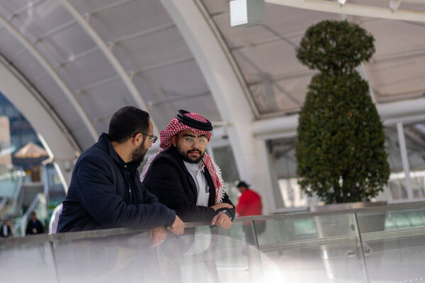 Two arabic males are talking to each other in modern place outdoors like mall shop and huge playing areas about there work life and problems