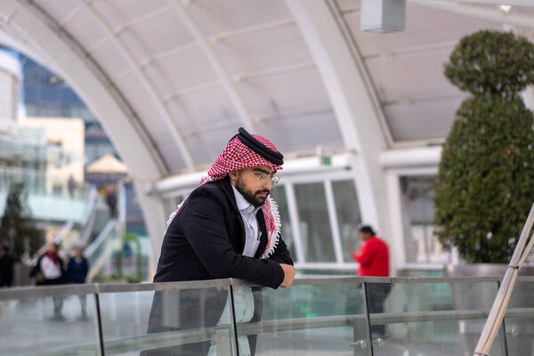 a stylish young man wearing a traditional Arabic headscarf and glasses, leaning against a glass railing in an urban setting. He exudes confidence and contemplation as he gazes into the distance