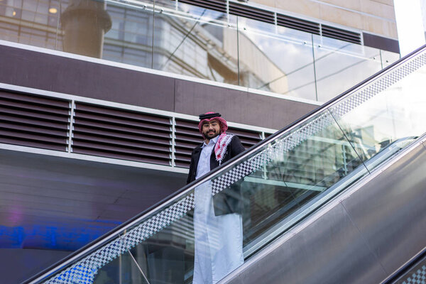 In the backdrop of a sleek, modern urban landscape, a young Arabic businessman stands confidently, surrounded by luxury cars and towering skyscrapers