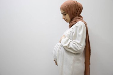 Ayrı beyaz arka planda hamile bir Arap kadın hamileliğin 9. ayında karnına dokunarak vücudunun şeklini gösteriyor.