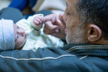 Büyükbaba yüzünde gülümsemeyle bebeği tutuyor, gri sakal, elinde bebek parmağı.