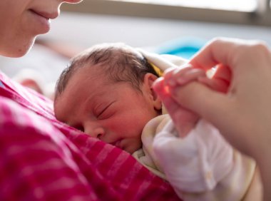 Anne yeni doğmuş bebeğini kucaklıyor. Yüzlerinde gülümseme ve bebek yüzünde sevimli bir ifade var.