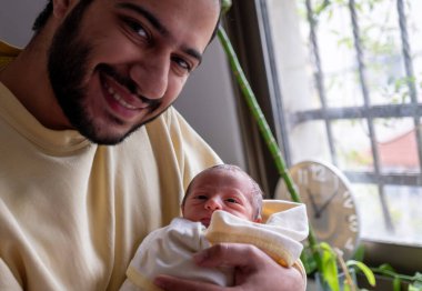 Baba yeni doğmuş oğlunu neşeyle kucaklıyor ve bebek yüzüne gülümsüyor.