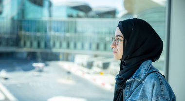 Pasaportunu havaalanında tutan genç Arap kadın arka planda uçakla yeni bir maceraya hazır.