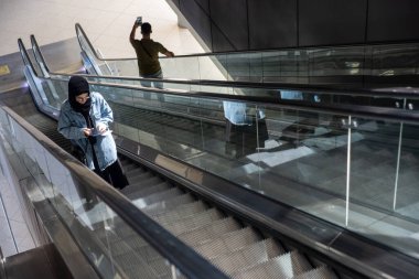 Havaalanında yürüyen merdiven kullanan genç bir kadın pasaportunu tutuyor ve seyahat sırasında cep telefonunu kullanıyor.