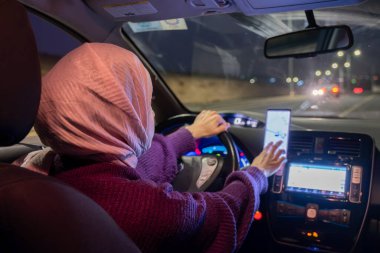 Gece araba süren Arap müslüman kadın kışlık giysiler, başörtüsü ve emniyet kemeri takıyor güvenli bir şekilde giriyor ve cep telefonu ve GPS ile haritalar kullanıyor.