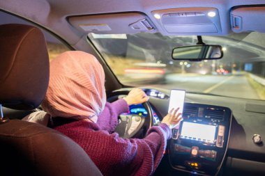 Gece araba süren Arap müslüman kadın kışlık giysiler, başörtüsü ve emniyet kemeri takıyor güvenli bir şekilde giriyor ve cep telefonu ve GPS ile haritalar kullanıyor.