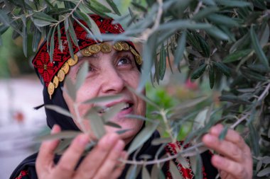 Zeytin ağaçlarında geleneksel giysiler giymiş bir kadının portresi elinde bir dalla yüzünde gülümsemeyle
