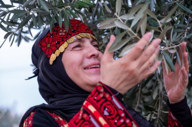 Zeytin ağaçlarında geleneksel giysiler giymiş bir kadının portresi elinde bir dalla yüzünde gülümsemeyle