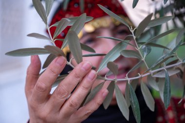 Zeytin ağaçlarında geleneksel giysiler giymiş bir kadının portresi elinde bir dalla yüzünde gülümsemeyle