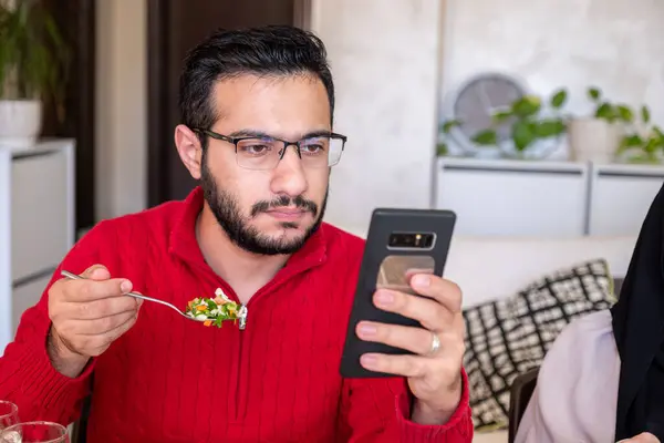 stock image Young bearded man using smartphone while eating