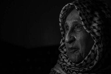 Karanlık arka planda beyaz Filistin keffiyesi giymiş, yüzünde gülümseme, özgürlük ve Filistinliler için dönüş hakkı arayan yaşlı bir kadının portresi.
