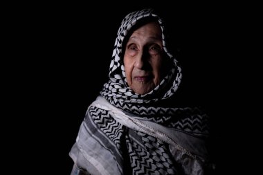 Karanlık arka planda beyaz Filistin keffiyesi giymiş, yüzünde gülümseme, özgürlük ve Filistinliler için dönüş hakkı arayan yaşlı bir kadının portresi.