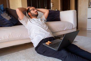 Sakallı erkek, evinden bilgisayarına bakarken kahve içiyor ve kurabiye yiyor.