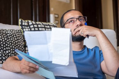 Oturma odasında oturan genç erkek makbuzları kontrol ediyor ve hesap makinesi kullanarak yaşam masraflarını hesaplıyor.