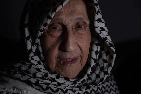 stock image portrait of an old lady in dark background wearing white palestinian keffiyeh with smile on her face looking for freedom and hope to Right of Return for Palestinians