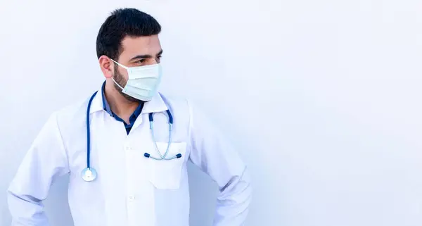 Genç erkek, maske takıyor ve doktor önlüğü giyiyor. Steteskopu kendine güvenen bir şekilde boynunda tutuyor.