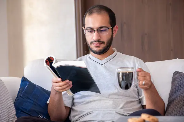 stock image Bearded arab man drinking coffee while reading book and sitting on living room couch