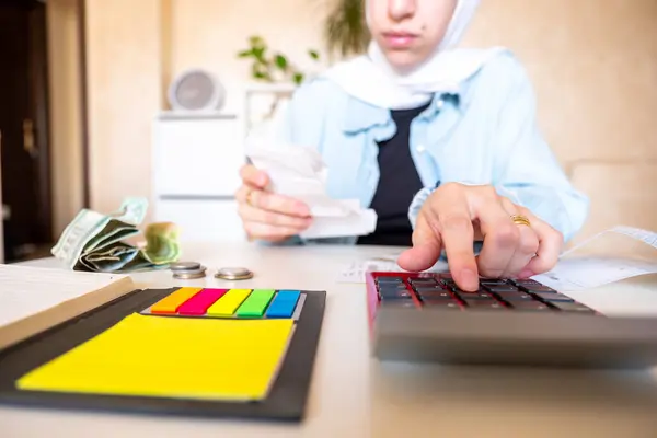 Oturma odasında oturan tesettürlü genç bir kadın makbuzları kontrol ediyor ve yaşam masraflarını hesaplamak için hesap makinesi kullanıyor.