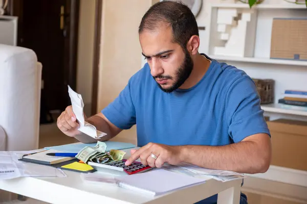 Genç sakallı erkek oturma odasında yaşam masraflarını hesaplıyor.