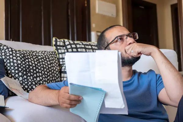 Oturma odasında oturan genç erkek makbuzları kontrol ediyor ve hesap makinesi kullanarak yaşam masraflarını hesaplıyor.