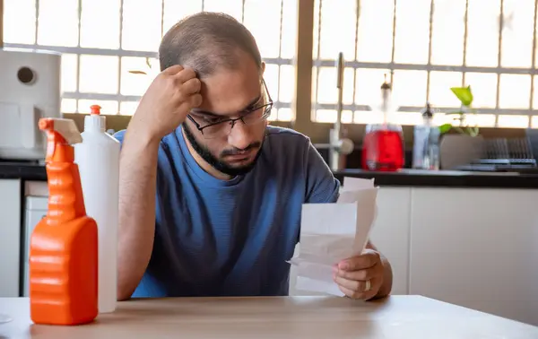 stock image man sitting in the kitchen holding receipt feeling frustrated due inflation and increase of daily needs supplies for house with detergents in front of him