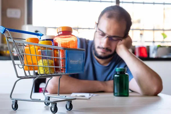 stock image man sitting in the kitchen feeling frustrated due inflation and increase of daily needs supplies for house with shopping cart in front of him holding medicine