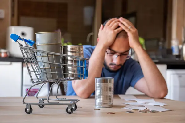 stock image man sitting in the kitchen holding receipt feeling frustrated due inflation and increase of daily needs supplies for house with shopping cart in front of him holding food