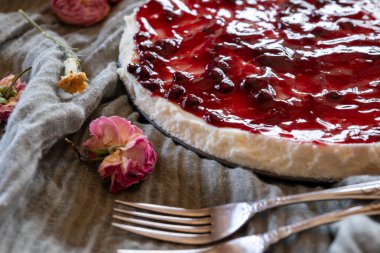 Çevresinde kiraz jölesi ve güller olan soğuk bir cheesecake, ahşap arka planda servis edilir. Yeşil fırlatma ve antika çatallarla.