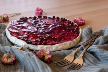 Çevresinde kiraz jölesi ve güller olan soğuk bir cheesecake, ahşap arka planda servis edilir. Yeşil fırlatma ve antika çatallarla.