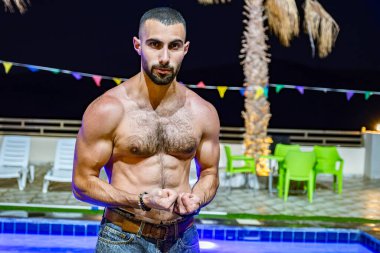 Genç sakallı erkek tatil boyunca havuz önünde kaslarını gösteriyor. Kendine güvenen bir yüz ve poz veriyor.