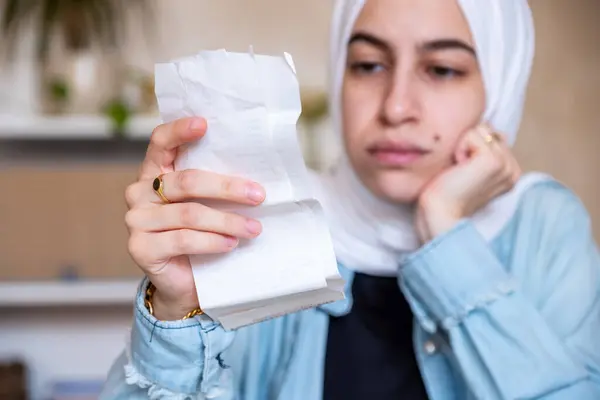 Oturma odasında oturan tesettürlü genç bir kadın makbuzları kontrol ediyor ve yaşam masraflarını hesaplamak için hesap makinesi kullanıyor.