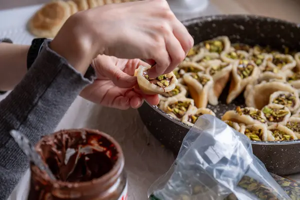 stock image Hands holding Qatayef filled with chocolate and topped with pistachios on a wooden table, with a plate to be prepared in the oven later as Ramadan sweets.