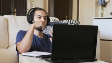 Genç bir adam oturma odasında oturuyor, uzaktan bilgisayarla çalışıyor. Kulaklık takıyor, internetteki bir webinara katılıyor ve not defterine yazıyor. Yanında bir evcil hayvan, serbest ve e-öğrenimi dengeliyor.