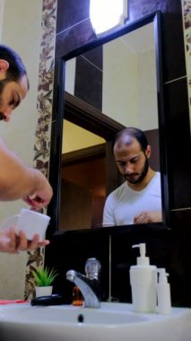 Orta yaşlı erkek, sabah sabah banyoda beyaz tişört giyerek kulak kıracağı kullanıyor.