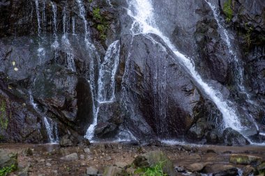 Sırt çantası ve açık hava kıyafetleriyle çağlayan bir şelalenin yanında duran ve doğa yürüyüşünün tadını çıkaran gülümseyen bir adam.